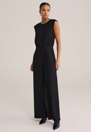 Femme debout portant une combinaison sans manches noire avec des jambes larges, des escarpins noirs pointus, des boucles d'oreilles en argent et un bracelet en argent.