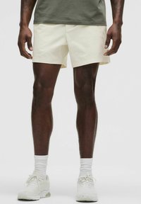 Lyse gule shorts med en afslappet pasform, lavet af blødt stof. Iført med hvide sneakers og hvide ankelstrømper, båret af en person stående.