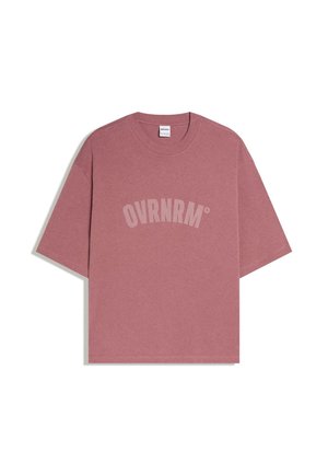 Poros rózsaszín, túlméretezett, rövid ujjú póló kerek nyakkal, elöl nagy, ívelt világos rózsaszín betűkkel nyomtatott "OVRNRM" felirattal.