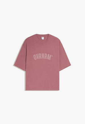 Prava roza velika kratka majica s kratkimi rokavi in okroglim ovratnikom, na sprednji strani je natisnjen napis "OVRNRM" v velikih, ukrivljenih svetlo roza črkah.