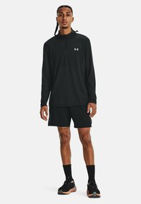 Sort langærmet atletisk top med lynlås og logo, parret med sorte shorts og sorte sneakers. Strukturstof, afslappet pasform.