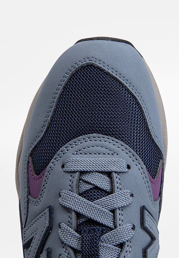 Zapatilla gris con parte superior de malla y acentos en azul marino, además de un acento morado en el lateral. Diseñada con cordones redondos y una puntera texturizada.