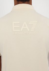 Krémové polo tričko s embosovaným logem EA7 a nápisem "Emporio Armani" pod ním. Hladký materiál, klasický design límečku.