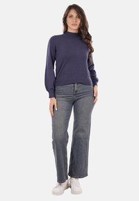 Pullover blu navy con collo alto, maniche a palloncino, polsini a coste e una texture liscia, abbinato a jeans grigi a vita alta e sneakers bianche.