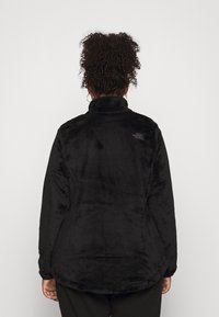 Persona con capelli ricci che indossa una giacca in pile nera, in piedi e girata di spalle contro uno sfondo chiaro e semplice.