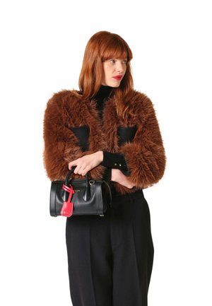 Femme aux cheveux roux portant une veste en fausse fourrure marron et un pantalon noir, tenant un sac à main noir avec une étiquette rouge, regardant sur le côté.