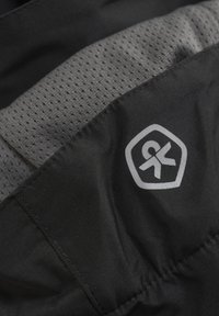 Svart tyg med en jämn textur och grå nätdetalj. Har en hexagonal logotyp i grått, placerad på sidan.