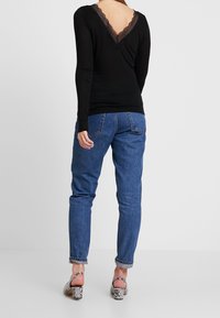 Černý top s dlouhými rukávy a krajkovým výstřihem do V, kombinovaný s modrými džíny a vzorovanými sandály na podpatku. Zadní pohled ukazuje detaily na zadní části topu.