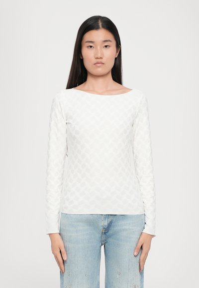T-shirt blanc à manches longues avec un motif texturé géométrique, doté d'un col large et associé à un jeans bleu clair déchiré.