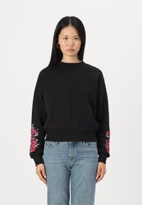 Μαύρη μπλούζα με μακριά μανίκια, διακοσμημένη με πολύχρωμο κεντητό λουλούδι στο αριστερό μανίκι. Φτιαγμένη από υφ�ασμάτινη δομή, άνετη εφαρμογή.