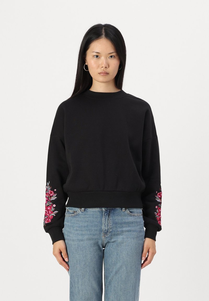 Μαύρη μπλούζα με μακριά μανίκια, διακοσμημένη με πολύχρωμο κεντητό λουλούδι στο αριστερό μανίκι. Φτιαγμένη από υφασμάτινη δομή, άνετη εφαρμογή.