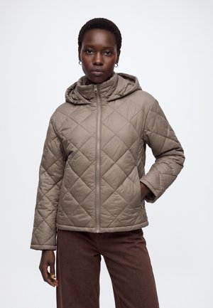 Femme portant une veste matelassée taupe avec capuche, debout, une main dans la poche, devant un fond clair uni.