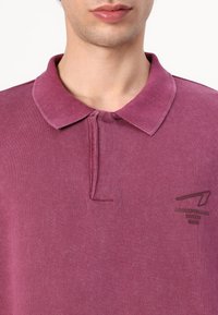 Polo en tissu rose foncé avec col et patte de boutonnage à trois boutons. Présente un petit logo sur le côté gauche. Texture douce.