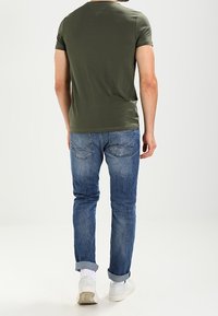T-shirt aderente color verde oliva abbinato a jeans in denim blu. I jeans hanno un orlo arrotolato e le scarpe sono sneakers bianche.