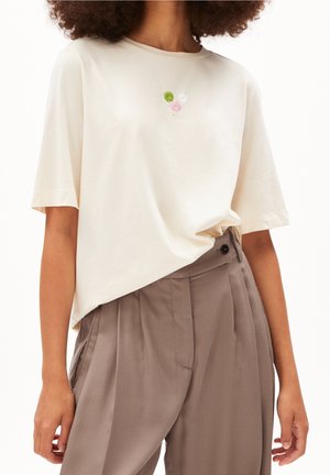 Kobieta ubrana w kremowy T-shirt z krótkim rękawem z małą kwiatową haftem oraz wysokim stanem taupe plisowane spodnie.