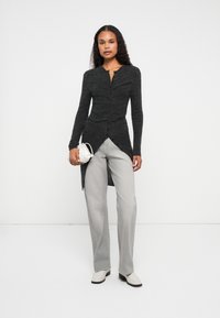 Cardigan longo cinza com fecho de botão e detalhes franzidos. Usado sobre calças largas cinza claro, complementado por uma pequena bolsa branca.