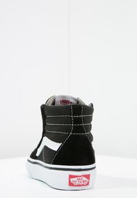 Czarne zamszowe sneakersy high-top z białymi skórzanymi akcentami, wyłożoną kołnierz i czerwonym logo Vans na pięcie. Posiadają białe przeszycia oraz teksturowaną powierzchnię.