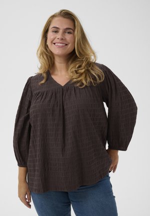 Lächelnde Frau mit langen blonden Haaren, die eine lockere dunkelbraune texturierte Bluse und blaue Jeans trägt, vor einfarbigem Hintergrund stehend.