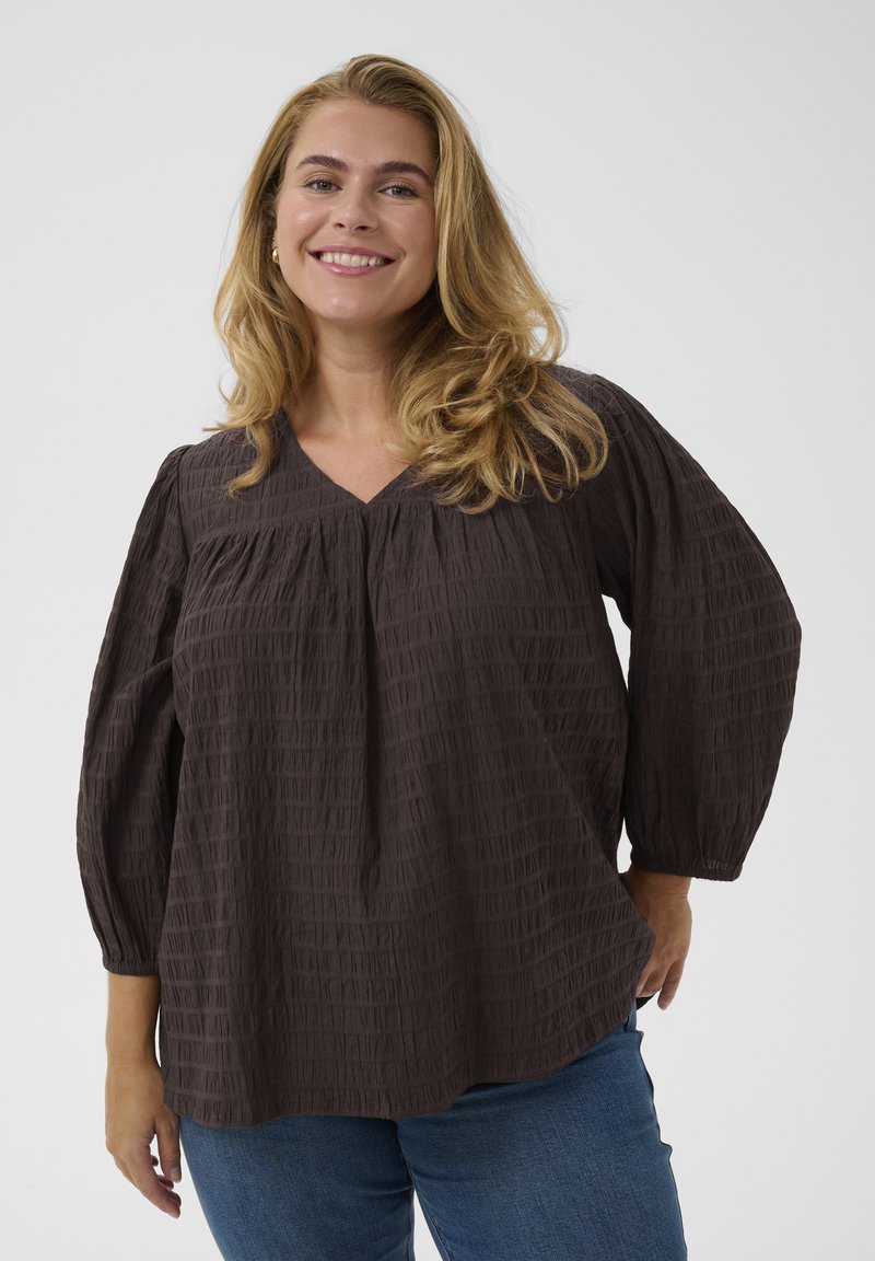 Femme souriante aux longs cheveux blonds portant un chemisier ample marron foncé texturé et un jean bleu, debout devant un fond uni.