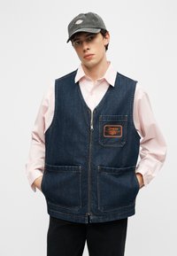 Νεαρός άνδρας που φοράει ένα τζιν γιλέκο με φερμουάρ πάνω από ένα απαλό ροζ πουκάμισο και ένα σκούρο γκρι καπέλο, στέκεται με τα χέρια στις τσέπες του γιλέκου.