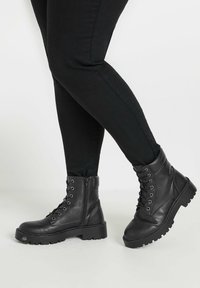 Botas de tobillo de cuero negro con puntas redondas, parte delantera con cordones, cremallera lateral, suelas de goma gruesas y detalles de costura reforzada.