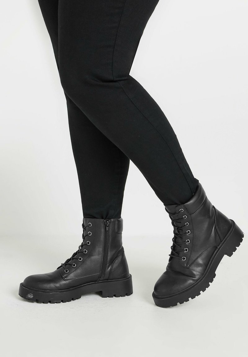 Botas de tobillo de cuero negro con puntas redondas, parte delantera con cordones, cremallera lateral, suelas de goma gruesas y detalles de costura reforzada.