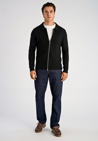 Czarny rozpinany kardigan z kołnierzem, o dzianinowej fakturze i ściągaczowych mankietach, zestawiony z ciemnymi dżinsami i brązowymi butami.