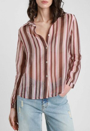 Donna che indossa una camicia trasparente a maniche lunghe con bottoni, a righe verticali marroni, rosa e bianche, abbinata a jeans azzurri consumati.