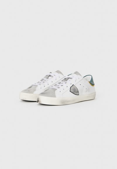 White sneakers with gray suede accents, rounded toe, flat sole, and contrasting green and yellow heel details. Lace-up design with perforations.