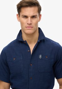 Camisa de manga corta azul marino hecha de tela texturizada, con un cierre frontal de botones, dos bolsillos en el pecho y un parche con el logo en el bolsillo izquierdo.