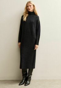 Blonde vrouw die een lange zwarte coltrui gebreide jurk en zwarte leren laarzen met puntige neus draagt, staand tegen een effen beige muur.