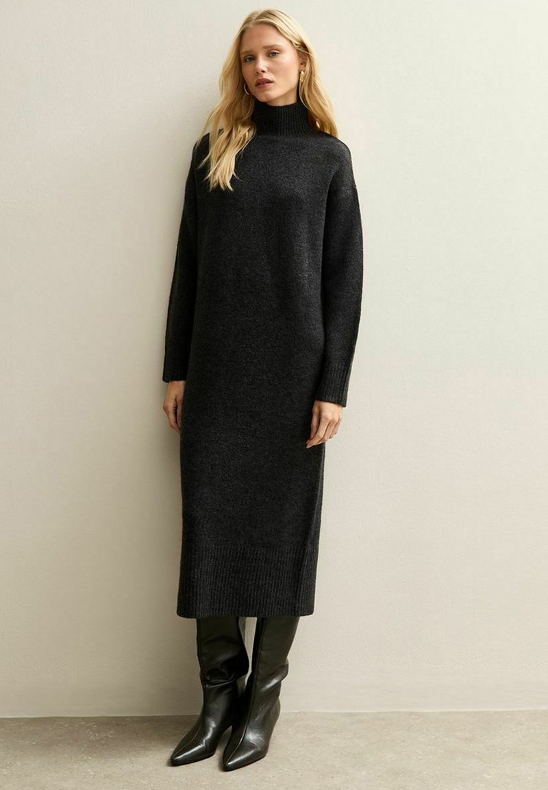 Blonde vrouw die een lange zwarte coltrui gebreide jurk en zwarte leren laarzen met puntige neus draagt, staand tegen een effen beige muur.