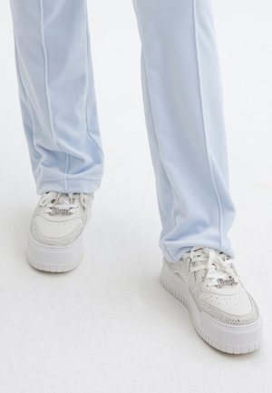 Sneakers bianchi con accenti argentati e strass, abbinate a pantaloni sportivi azzurri chiari. Presentano una suola spessa e un design con lacci.