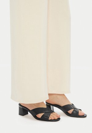 Schwarze Leder-Sandalen mit Absatz und Kreuzriemen, quadratischer Spitze und mittelhohem Blockabsatz. Getragen zu weiten cremefarbenen Hosen.