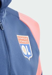 Niebieska sportowa kurtka z różowymi akcentami na ramionach; posiada haftowane logo Olympique Lyonnais oraz zamek błyskawiczny z przodu. Miękka tekstura materiału.