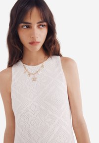 Top in pizzo bianco con motivo a diamante, caratterizzato da uno scollo alto e un tessuto testurizzato. Collana in oro con charm e dettagli.