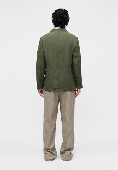 Άτομο με κατσαρά μαλλιά που φοράει μια ελιά πράσινο μπουφάν και φαρδιά μπεζ παντελόνια, στέκεται με την πλάτη προς ένα απλό φωτεινό φόντο.