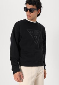 La sudadera negra presenta un gráfico de logo triangular en un tono ligeramente más claro, con puños acanalados y un diseño de cuello redondo.