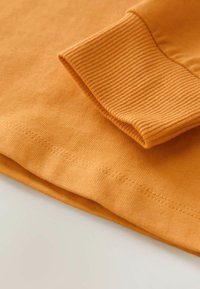 Oranžest puuvillane T-särk, millel on ribakujuline mansett ja lapik õmblusdetail. Kanga pehme tekstuur, sile ja ühtlane viimistlus.