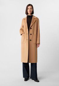 Donna con un lungo cappotto cammello sopra una maglia nera e pantaloni larghi scuri, in piedi contro uno sfondo bianco semplice.
