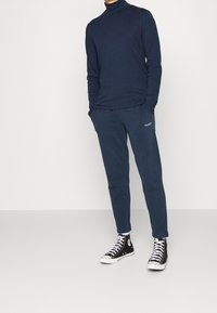 Tummansininen pitkähihainen poolopaita, yhdistettynä samansävyisiin tummansinisiin jogger-housuihin. Mustat korkeavartiset lenkkarit valkoisilla yksityiskohdilla. Yksinkertainen, rento muotoilu.