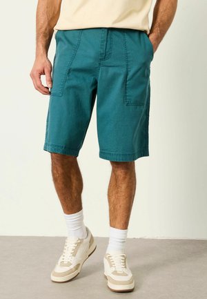 Mann trägt türkisfarbene knielange Shorts mit Fronttaschen, weiße Socken und beige-weiße Sneakers, steht vor hellem Hintergrund.