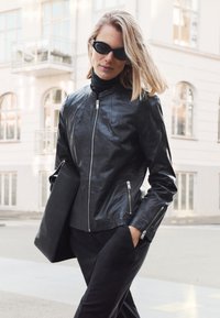 Kvinde iført sort læderjakke og solbriller, bærende på en sort mappe, gående på en byvej med lyse farvede bygninger i baggrunden.