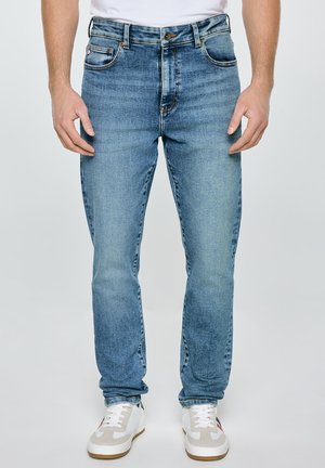 Mężczyzna w jasnoniebieskich dżinsach i białych trampkach, stoi na jednolitym tle.