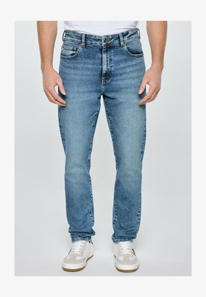 Mężczyzna w jasnoniebieskich dżinsach i białych trampkach, stoi na jednolitym tle.