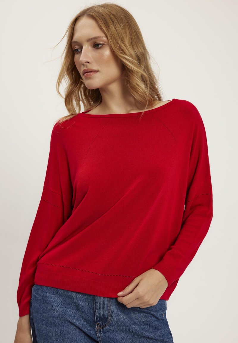 Czerwony sweter dzianinowy z szerokim dekoltem, opadającymi ramionami i ozdobnym przeszyciem wzdłuż rękawów. Połączony z niebieskimi dżinsami.
