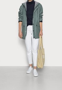 Henkilö, jolla on päällään vihreä sadejacke, musta poolopaita, valkoiset housut, valkoiset lenkkarit ja joka pitää kädessään beigeä kangaslaukkua, jossa on teksti kierrättämisestä.