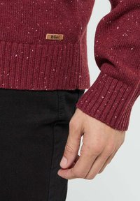 Main droite détendue à côté d'un pantalon noir, portant un pull en maille bordeaux avec des éclaboussures blanches et une petite étiquette marron Lee Cooper sur le devant.