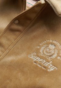 Lichtbruin corduroy shirt met ritsluiting, met witte geborduurde merktekens en een subtiele ribbelstructuur.