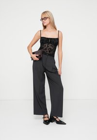 Top in pizzo floreale nero con spalline sottili, abbinato a pantaloni grigi a vita alta e a gamba larga. Ballerine nere a punta e una pochette marrone completano il look.
